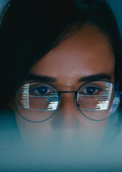 Um close-up dos olhos de uma pessoa a usar óculos, refletindo código e dados que brilham em azul.