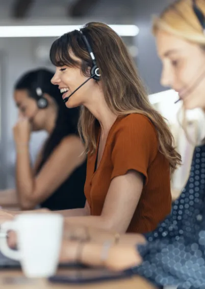 Uma mulher a sorrir a usar auscultadores com microfone numa secretária, com colegas de trabalho ao fundo em um escritório.