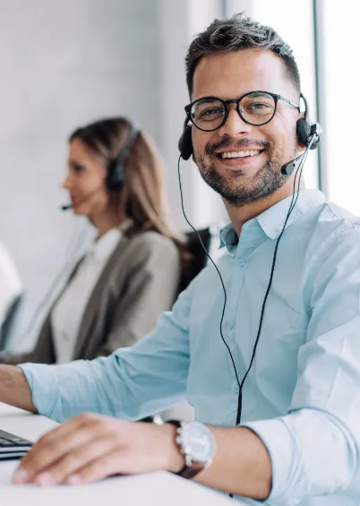 Um homem de camisa azul, a sorrir para a câmara, a usar óculos e auscultadores com microfone, enquanto uma colega está desfocada ao fundo.
