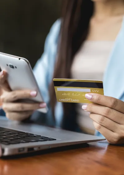 Uma pessoa a segurar um telemóvel e um cartão de crédito, com um computador portátil em primeiro plano, a representar compras online.