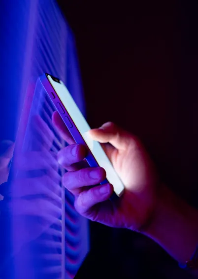 Uma mão a segurar um smartphone no escuro, com o ecrã a brilhar intensamente em tons de azul e roxo.