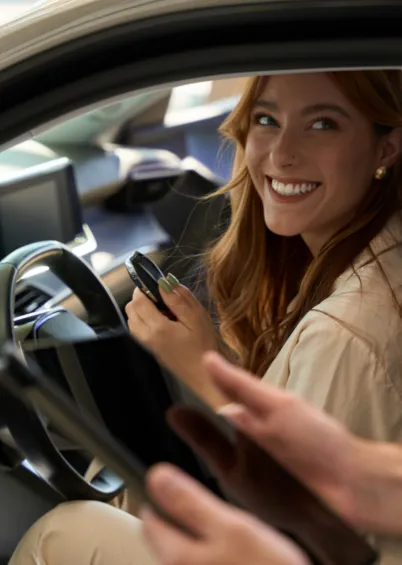 Uma mulher sorridente no banco do motorista de um carro, a segurar as chaves, enquanto outra pessoa, fora de foco, lhe entrega um tablet.