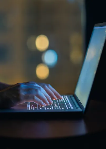 Mãos a digitar num teclado de computador portátil retroiluminado, com luzes de bokeh desfocadas ao fundo.